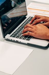 Hands typing on a laptop at a desk with an insurance paper and plant, suggesting a work environment.